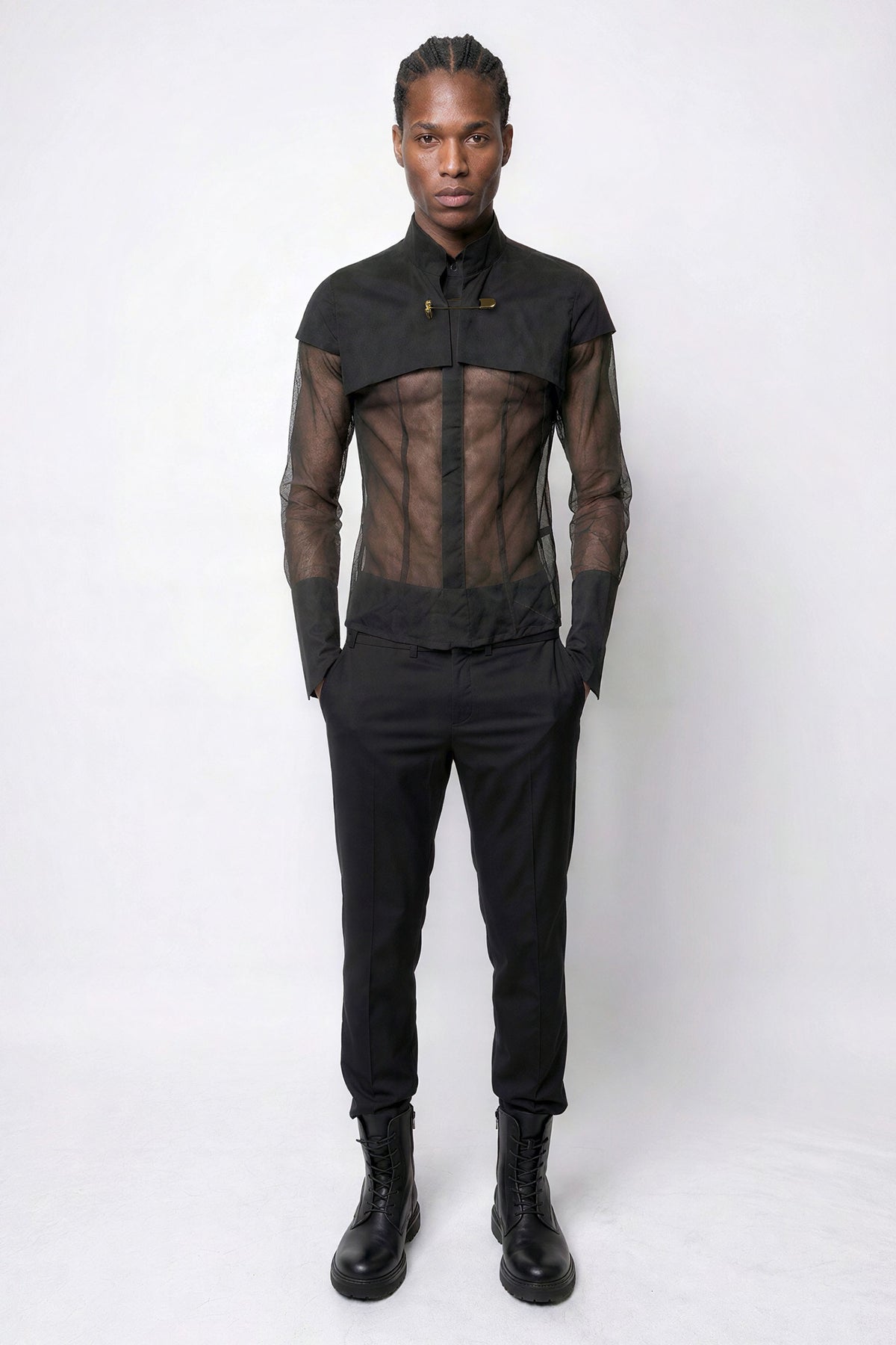 A model stands against a light gray studio background wearing a Esther Perbandt black sheer mesh long-sleeve shirt with the structured crop SUSPENSE Vest, black trousers, and black boots.