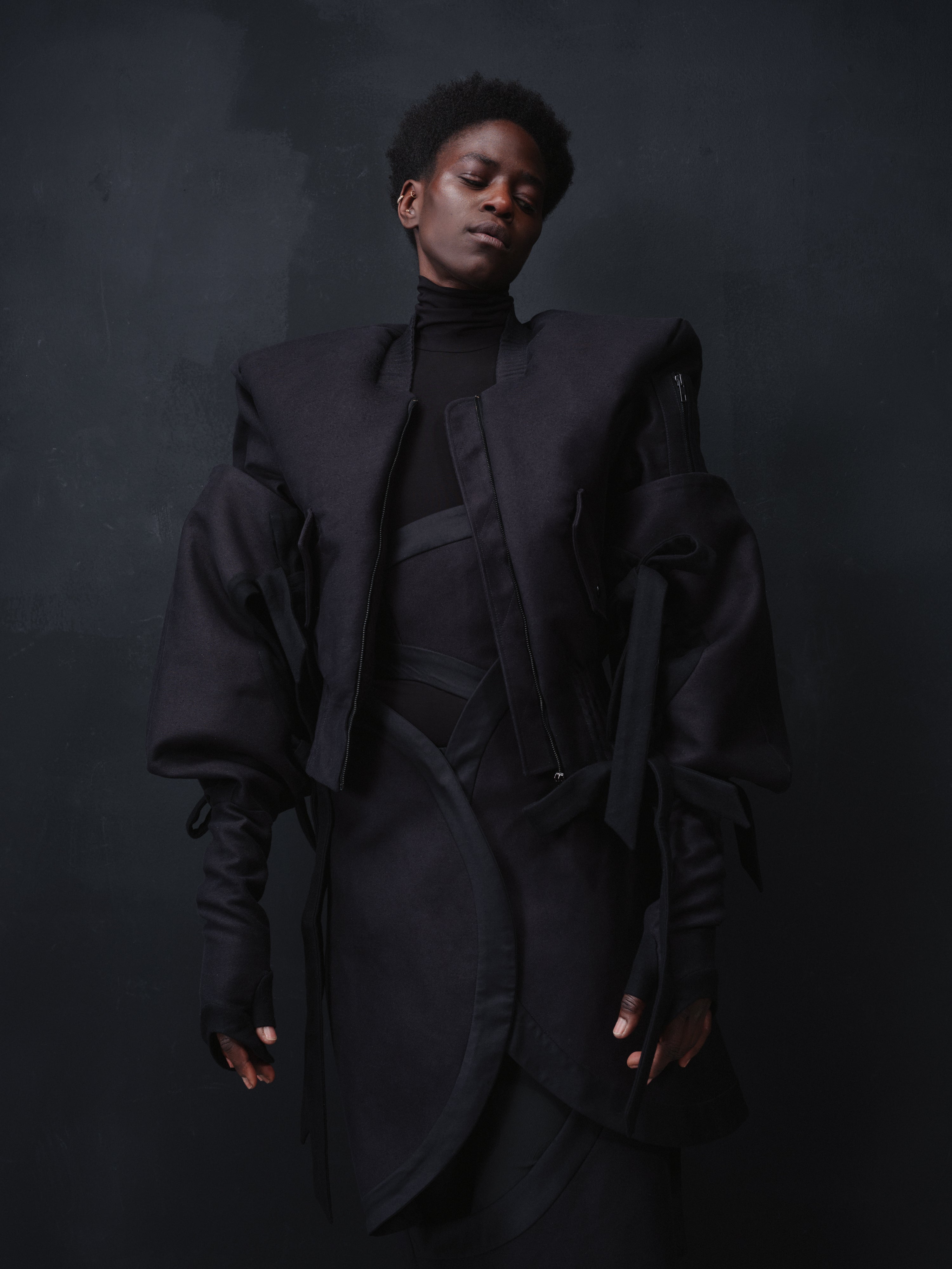 A model wearing a dark, futuristic black outerwear bomber jacket "Alles Von Hier" with dramatic voluminous sleeves and layered details over a black turtleneck, set against a dark textured studio background.
