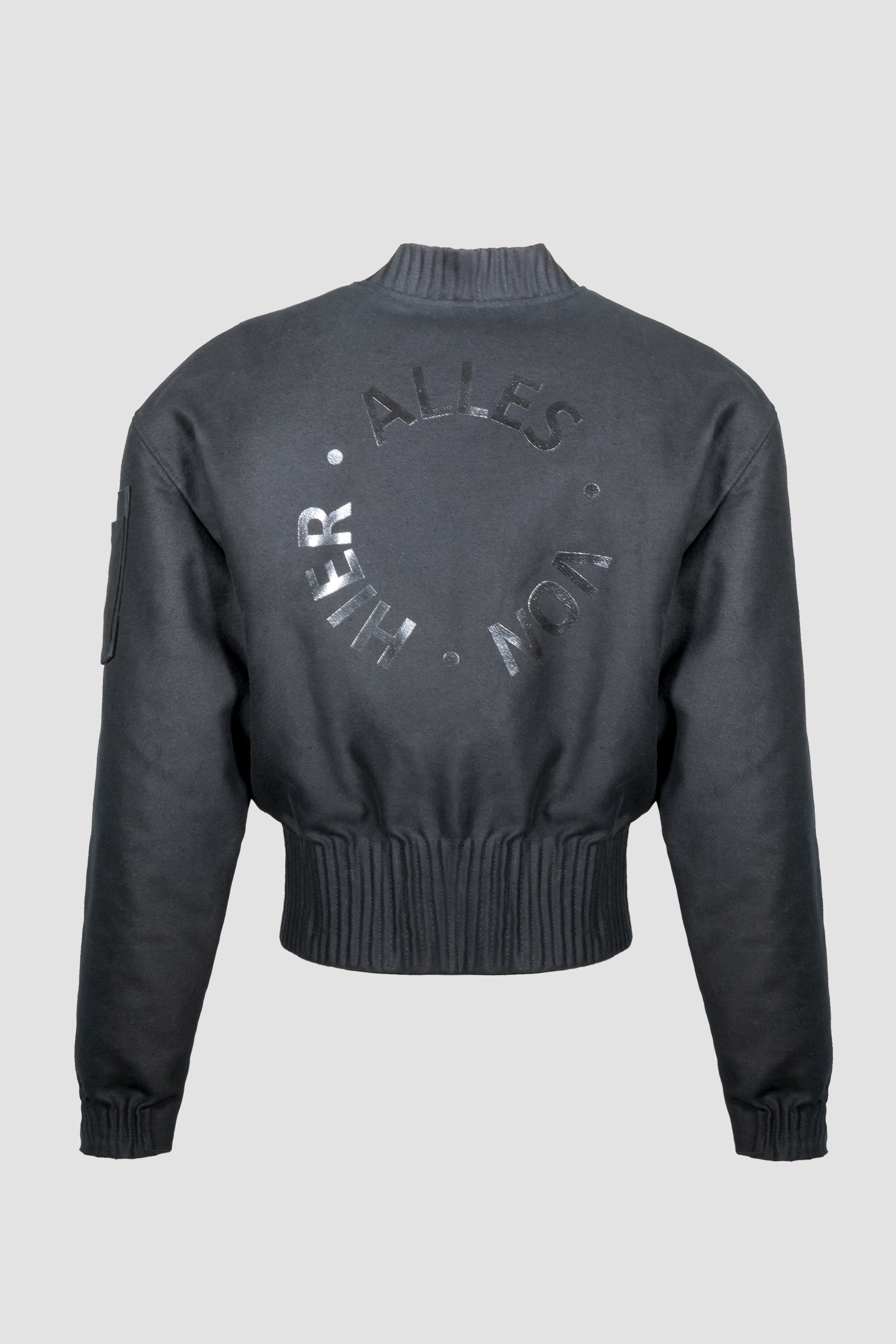 A black bomber jacket "Alles-Von-Hier" (Everything from Here) by Esther Perbandt, shown from the back, with a ribbed knit collar, cuffs, and hem, with a tonal circular logo reading. 