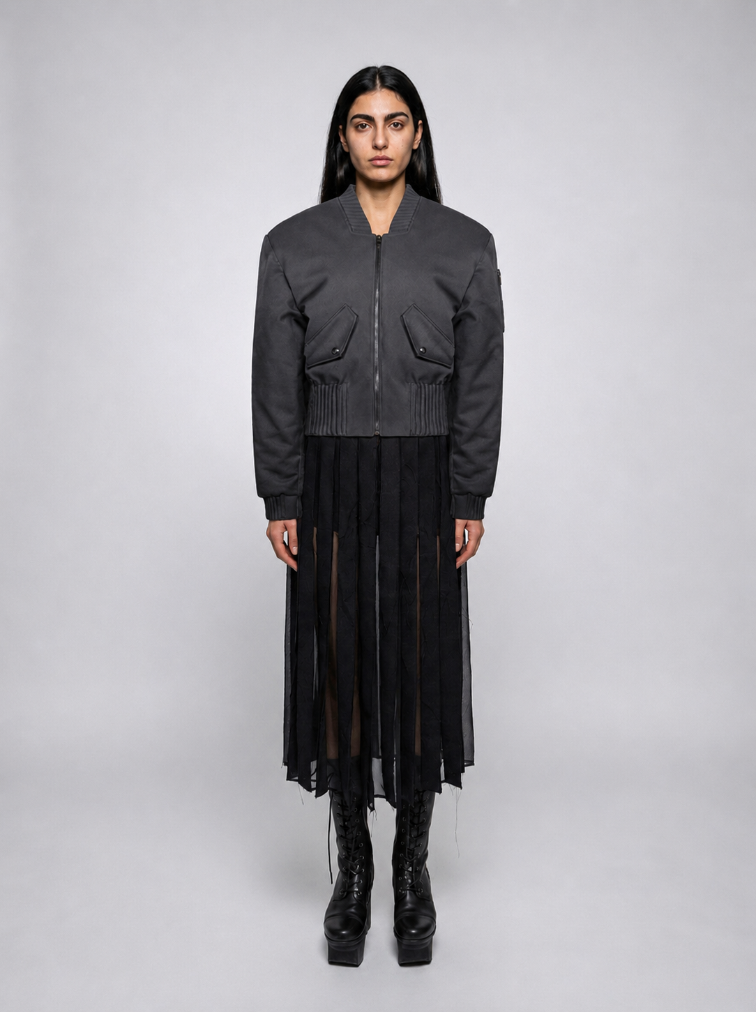 A model stands against a light studio background wearing a black bomber jacket "Alles Von Hier" (Everything from Here) by Esther Perbandt, with a front zipper and flap pockets, paired with a long black pleated mesh skirt and black platform boots.