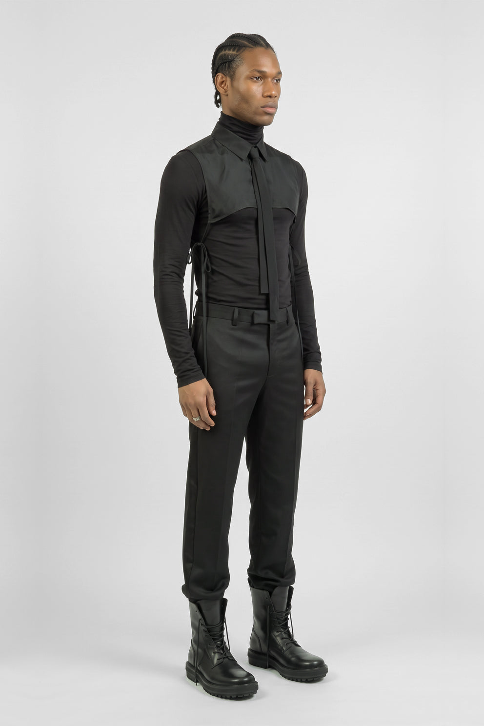 A model wearing a black futuristic full-body jumpsuit with a high collar, long sleeves, structured shoulder panels, side lacing, and matching black pants and combat boots, photographed against a light gray studio background.