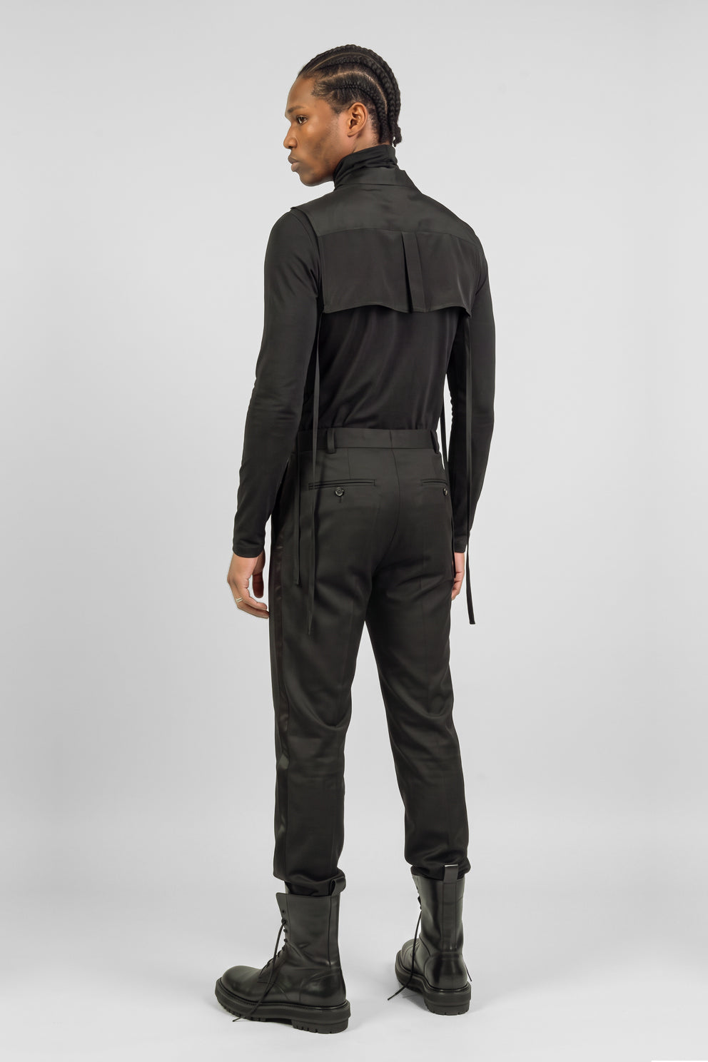A model shown from the back wearing a black long-sleeve high-collar top with a unique shoulder panel, black trousers, and black combat boots, set against a plain light gray studio background.