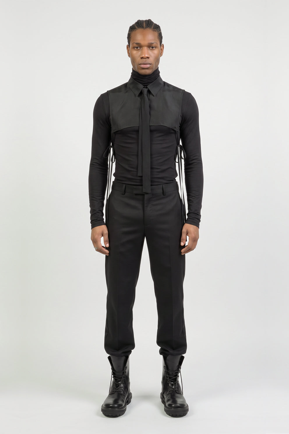 A model stands facing forward in a black long-sleeve turtleneck jumpsuit with a tie collar and side lacing details, paired with black boots, against a light gray studio background.