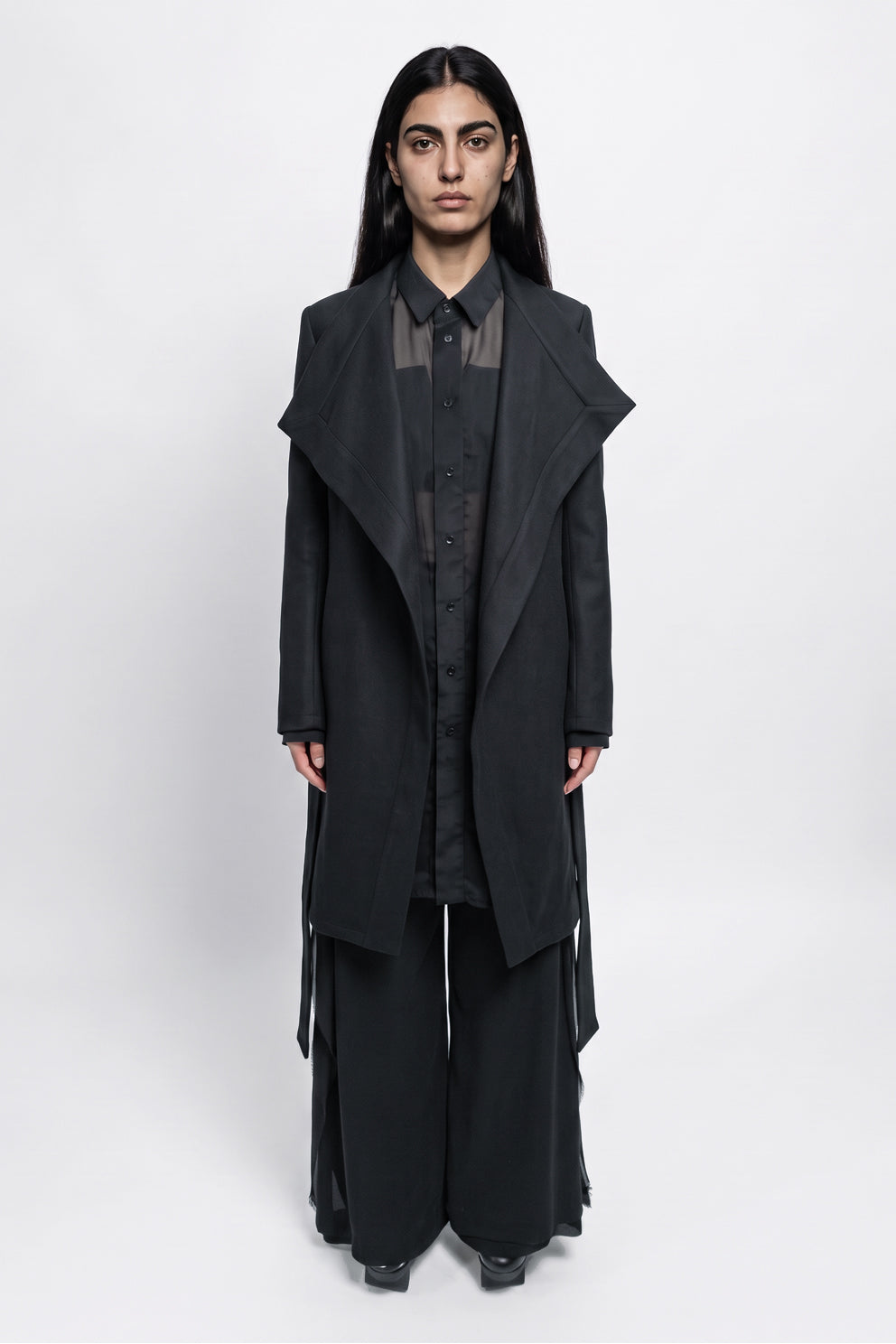 A model stands facing forward in a studio wearing a long black asymmetrical coat over a black shirt and wide-leg black pants against a light gray background.