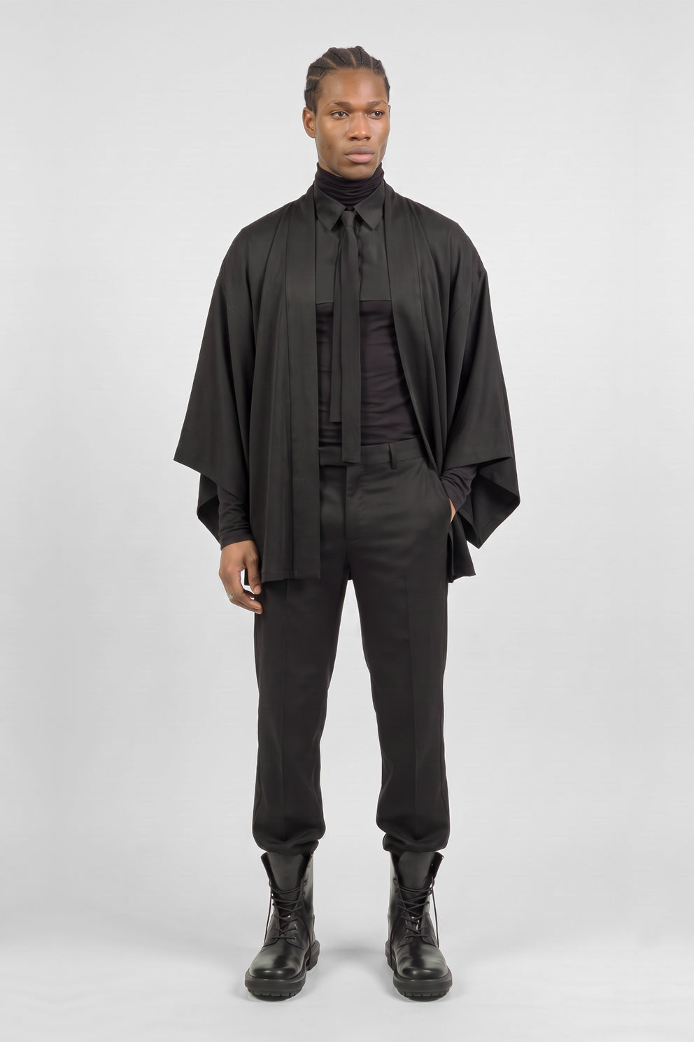 A model stands against a light gray studio background wearing a full black outfit consisting of a kimono, a turtleneck, the wild overshirt tied, slim-fit trousers, and black Tarone boots.
