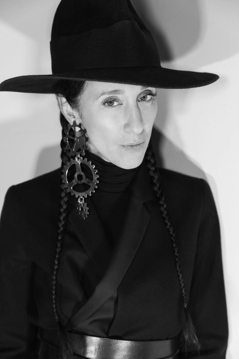 Portrait of Esther Perbandt wearing a wide-brimmed black hat, an oversized triple wheel gold earring, and dark clothing with decorative elements.