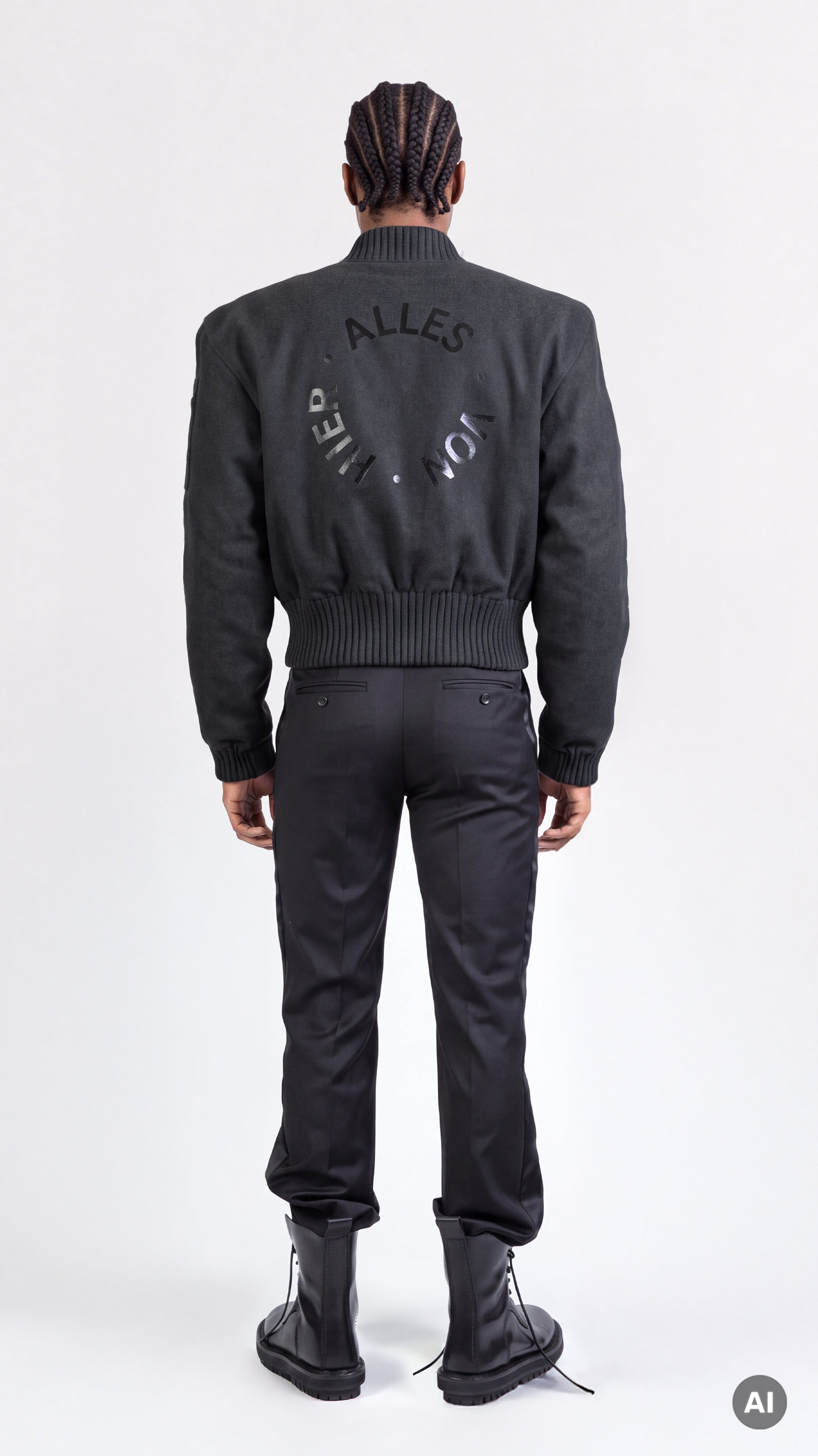 A model shown from the back wearing the black bomber jacket "Alles Von Hier" (Everything from Here) by Esther Perbandt, with a tonal circular graphic on the back, paired with black smoking black pants, and black trippen boots.