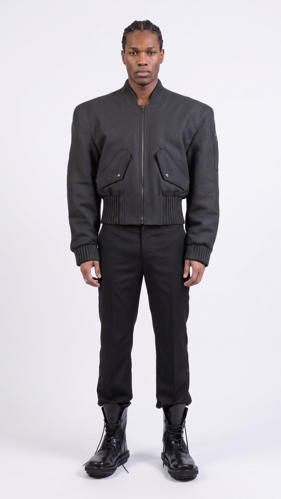 A model stands against a light gray studio background wearing a black bomber jacket "Alles Von Hier" (Everything from Here) by Esther Perbandt, with a front zipper and flap pockets, paired with black smoking black pants, and black trippen boots.
